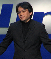 A Japanese man wearing a pair of glasses, a black suit jacket, and a black shirt, facing to the left. The man stands behind a blue and white background.