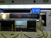 An airport check-in counter in black colour theme, with Japan Airlines written in capitals in white colour. There is a black carpet with the extra large letter 'F' in script font and 'JAL First Class' in smaller font, both in gold colour. The foreground has gold coloured poles with red velvet ropes