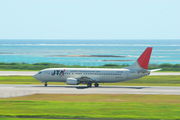 A Boeing 737-400 aircraft painted in Japan Transocean Air livery, travelling along the runway during take-off, with a clear green grass strip in the foreground and a blue sea view in the background