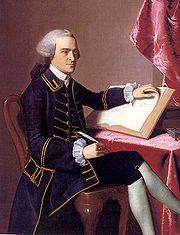 Full-length portrait of a young man seated at a table. He wears a finely tailored dark suit, knee breeches with white stockings, and a wig in the style of an English gentleman. He holds a quill pen in his right hand, and is turning the pages of a large book with the other hand.