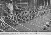 A row of more than a dozen children stretches into the distance.. They sit holding wooden looms with two pieces of thread leading from each. A man stands behind them in front of some wooden structures.