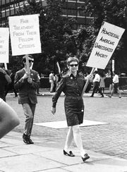 A black and white photograph of a woman holding a sign that reads "Homosexual Americans Unrecognized Minority"; she is wearing a dark dress, pumps, and wearing sunglasses. A man is behind her in a mod suit holding a sign that reads "Fair treatment from their fellow citizens". Following them both is a line of picketers circling a plaza far in the background