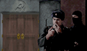 A building with two wooden doors and a hammer & sickle engraved above them, a Soviet soldier played by Frank Klepacki lighting a cigarette, a man dressed in black moving up behind him