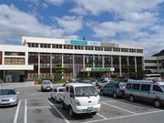 In front of a low and ivory building, a parking lot and cars are shown.