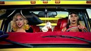 A blond woman sitting beside a brunette woman who is driving a yellow colored van. The blond woman wears a giant hat on her head. A pair of dice hangs from the rear-view mirror between them.