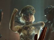 A blond woman wearing a khaki colored bustier and khaki cap with the word GAGA written on it. She looks towards the left of the image. Her left hand is folded in front of her and the right hand is gestured upwards. The silhouette of a male dancer is visible to her left.