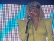 The bust image of a blond woman in a yellow dress with shoulder pads. Her hair is in bob cut and she wears video sunglasses. She holds a microphone in her right hand to her mouth. The backdrop video displays diagonal indigo and white stripes.