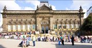 A queue of people at the edge of a pedestrianised square stretches uphill across the scene from right to left, then doubles back up another slope to the double-arched entrance at the centre of a large mid-nineteenth-century stone building.  This also has steps leading up to the entrance with four ornamental street-lamps in front.  On either side of the entrance are giant pilasters, two more of which can be seen, surmounted by urns, at the building's corners.  There are six large windows with shell tympanums and scroll-effect balcony rails between the entrance and the corners on each side, with five small circular windows above and between the tympanums.  Below on each side are basement windows, those on the left obscured because of the sloping site.  Above the entrance is a large arch and above that, in gold lettering, is "Leeds City Museum".  Higher up is another gold inscription, "Leeds Institute".  Above this is another, larger, tympanum with sculpture, set in a pediment with urns on either side.  Behind is a mansard roof.