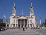 A pedestrian square in sunny weather.  At the rear is a large neo-Georgian building of Portland stone.  It has a giant portico with four Corinthian columns and a pediment.  On the ground floor, there are three entrance doors under the portico and two windows on either side of it.  All the doors and windows have Gibbs surrounds and pediments.  The window pediments are normal, while those over the doors are segmental.  The first floor has seven plain windows, three of them behind the portico, and all of them larger than those on the ground floor.  On either side of the building are two tall towers in Wren style with elaborate columned pavilions surmounted by smaller pedimented ones, and on top of each is an obelisk surmounted by a golden owl.  In front of the building on each side there are similar obelisks and owls, and prominent gold clocks are attached to the sides of the building at second-floor level.  In the background on both sides are tall modern buildings.