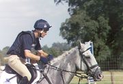 A gray horse, equipped in black tack, ridden by a man in a blue shirt and tan pants. In the background is a wire fence and trees.