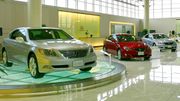 Car showroom displaying three sedans, the nearest on a glass turntable, in front of a reception counter and windows.