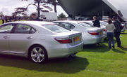 Five identical sedans lined up on a lawn. Two men and a woman are walking behind one sedan, carrying recording equipment.  The background has a stage, trailer, trees, and other persons.