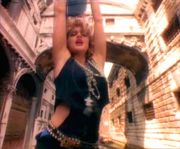 A blond woman with both her hands up in the air. The backdrop shows a number of houses with interconnecting channels. The woman wears a black top with a number of garlands around her neck, and a large chain-like belt. Her blond hair is curled and unkempt.