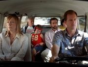 A film screenshot shows the family all seated in the Volkswagen Microbus as it is driven on a highway. The angle is from the windshield looking into the vehicle so that the majority of the interior can be seen. The mother is sitting in the front passenger seat with a dull expression on her face. The father is driving the vehicle with a smiliar expression. In the middle row, the daughter is looking down, listening to music from a CD player. Her uncle is seated on the right, looking to his left. In the back row, the grandfather is looking towards his grandson (who's face is slightly obscured by his father's head due to the angle). In the background, other vehicles can be seen driving on the highway.