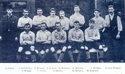 alt=A black-and-white photograph of a football team lining up before a match. Five players, wearing light shirts, dark shorts and socks are seated. Six more players immediately behind them. Two men in suits are standing on either side of the players.