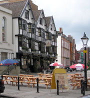 A seventeenth century timber framed building with three gables and a traditional inn sign showing a picture of a sailing barge. Some drinkers sit at benches outside on a cobbled street. Other old buildings are further down the street and in the background part of a modern office building can be seen.