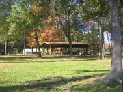 Leaves are turning red on trees in a park graced with a covered picnic shelter on a wide green lawn. Homes are visible on the far side of the park.