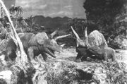 A black-and-white screenshot of three triceratops atop a cliff with trees surrounding them and mountains in the background.