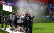 A crowd of men, some wearing grey suits and some wearing white shirts, navy shorts and white socks, celebrate raucously on a podium. An open bottle of champagne is visible in front of them, spiralling through the air as if somebody has thrown it.