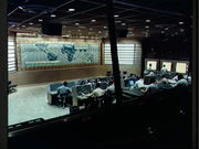 A large room seen through windows at the rear, with two rows of people seated at desks and computers, and a map taking up the whole of the front wall.