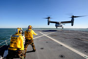  A V-22 performing a vertical landing on the USS New York with two of the ship's crew nearby.