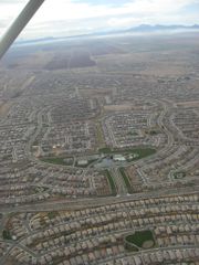 An aerial view of residential neighborhoods.  Homes line the curving roads that are placed within a larger grid of arterial streets.  Near the center is a large park with a pond.  Further in the background are undeveloped agricultural fields, and beyond them a range of mountains on he horizon.  A support strut from the airplane's wing is visible in the upper left corner.