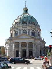 A building. It has a green dome, with four pillars for an entrance. Traffic is in the foreground.