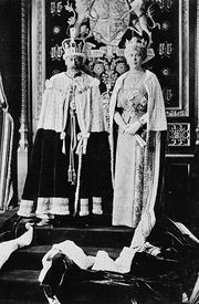 Late-middle-aged couple in crowns and ermine capes stand on a dais