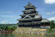 A  large 5-storied  castle tower with black wooden walls located on a platform of unhewn  stones surrounded on two sides by water. The tower is connected to lower  structure.