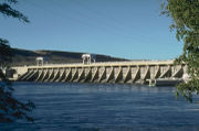 A concrete hydroelectric dam's spillway on the downriver side. Water is in the foreground and power lines run overhead.