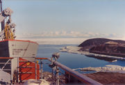  In left foreground are the upper works of a modern ship. To the right, on a low headland fringed with sea ice, isa square hut with a pitched roof. In te distance is a range of icy mountains