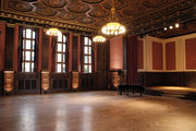 A ballroom with hardwood floors, and a ceiling decorated with chandeliers and octagon-shaped tiles. The wall on the left is decorated with dark wood panels, red walls, and windows. On the right is a small stage with a piano in front.