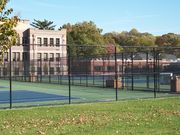 picture of a fence, with tennis courts behind them, with a three story brick building in the background.