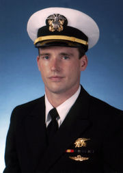 Color picture of Michael Murphy, a white male, wearing a military white dress hat and dark blue suit. There is a blue background behind him and he is wearing a gold Navy Seal Trident, 2 blue and green striped ribbons, 1 red and yellow striped ribbon and gold parachute insignia wings below the ribbons.