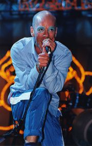 A middle-aged Caucasian male with a shaved head sings into a microphone while standing on some monitors. He is wearing a light blue button-up shirt with dark blue and white sweat pants and light blue makeup outlining his eyes. Behind him is a light display making the outline of a lotus flower.