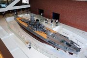 An overhead view of a very large warship model housed in a four-storey gallery. People are viewing the model and taking photographs.