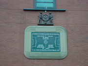 A stone tablet, inscribed with the image of a football pitch and several names. It is surrounded by a stone border in the shape of a football stadium. Above the tablet is a wooden carving of two men holding a large wreath.