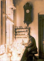 An old, bespectacled man wearing a suit and sitting at a bench by a large window. The bench is covered with small bottles and test tubes. On the wall behind him is a large old-fashioned clock below which are four small enclosed shelves on which sit many neatly labelled bottles.