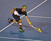 A dark-haired tennis player is reaching to hit a tennis shot with a racket in his left hand, and he is wearing black shoes and shorts with black and white mixture shirt and yellowish-green accessories