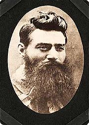 Head of a young man with a long, untrimmed beard, and with hair cropped above the ears, but longer and slicked strikingly up and back on the top. His moustache and beard are so long that his mouth and shirt front can barely be seen. His eyes look over the viewer's right shoulder .