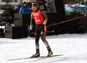 A woman in black winter sportswear stands on cross country ski in an upright position. She wears a red jersey with the number 5 and holds her ski poles horizontally behind her.