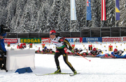 A woman on cross country skies wearing a red cap, a white jersey with the number 15 and black trousers skies away from a shooting range covered in snow. Half a dozen people in the background are shooting while lying on the ground.