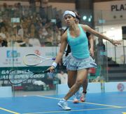 An indoor squash-court; a female player in light blue sportswear, a short pleated skirt, balancing for a shot, facing the camera and mostly obscuring her partner who is behind her. Both their rackets are at knee level on their left. A crowd is in the background, behind glass.