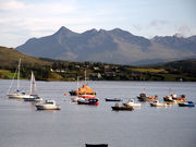 A body of water in the foreground contains several sailing vessels, including yachts, small fishing boats and an orange  lifeboat. In the middle distance a variety of modern cottages are set amongst coniferous trees on a long grassy slope. Large and precipitous black mountains dominate the background.