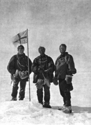  Three men stand around a flag planted in the snow.