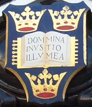 A shield displaying a coat of arms; on a dark blue background, an open book displays the words "Dominus Illuminatio Mea"; two gold crowns above, one below