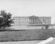 Massive, rectangular, three storied, granite palace with a flat roof; front pediment has six large columns. A large staircase leads to area with balustrades and to the front entrance with five rounded arches.