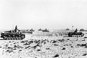 Several tanks advance to the right across the brush-covered desert.