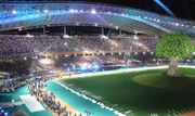 A portion of the stadium with stands full of people, a large artificial tree is on the right side of the image.  A group of people are walking together on the stadium floor