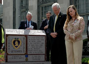 A color picture of Daniel and Maureen Murphy standing next to a monument in front of the Michael P. Murphy Post Office in Patchogue, New York. The monument has a purple heart and some wording inscribed on it and there are 2 men in the background.